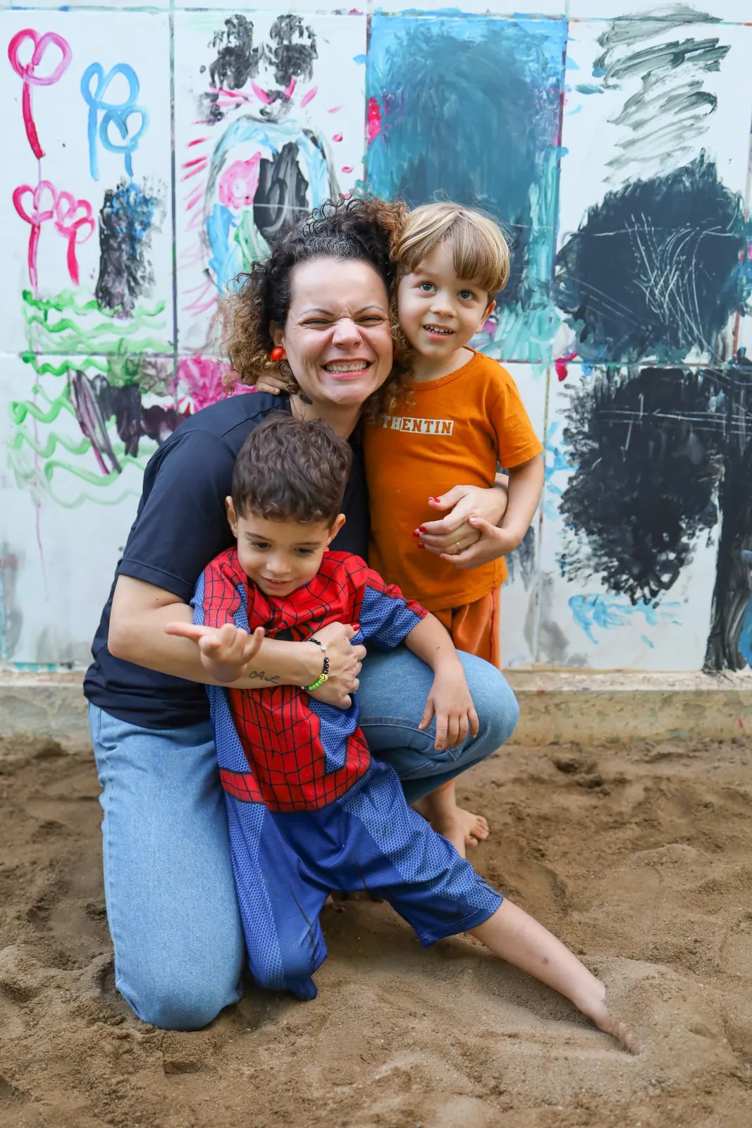 Educadora abraçada a duas crianças diante de um mural pintado