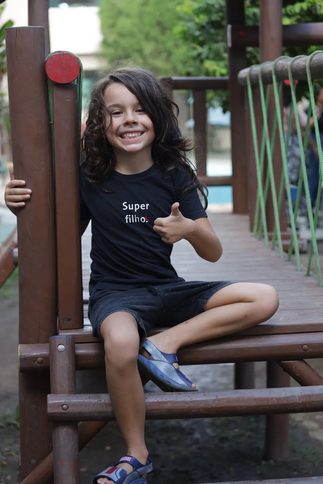 Menino sorri e faz sinal de positivo sentado no parquinho